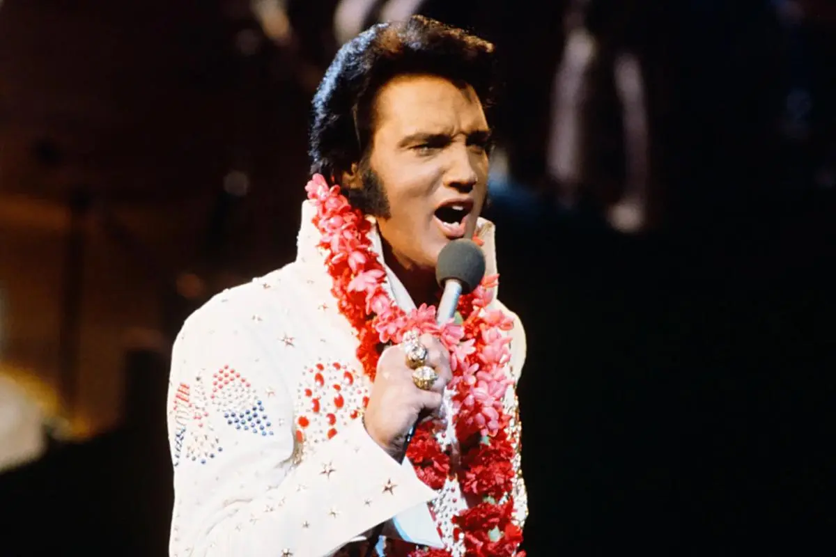 Elves Presley wearing a white jumpsuit and flower lei, singing into a microphone onstage.
