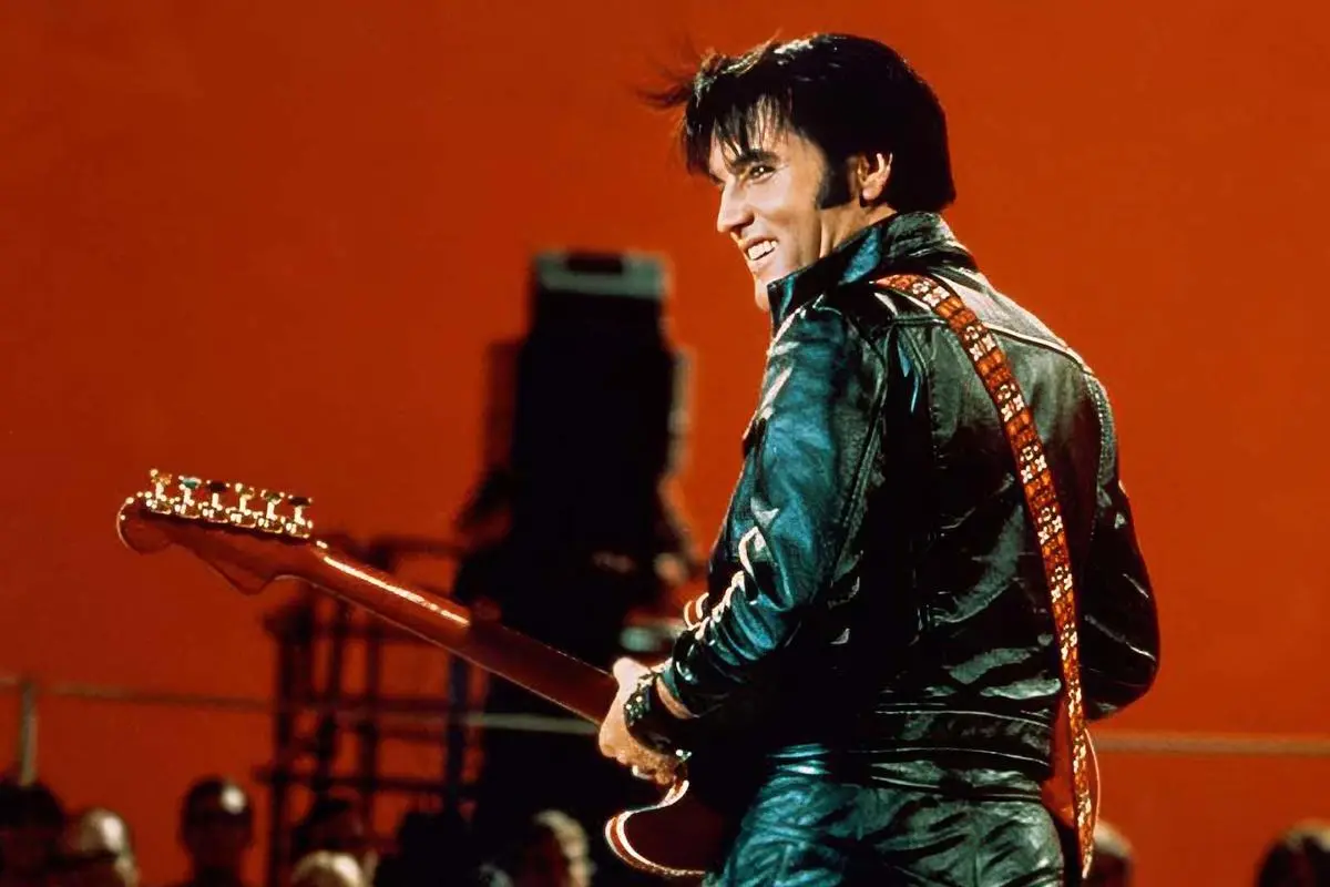 Elves Presley in a dark leather outfit playing electric guitar, turning toward the audience under stage lights.