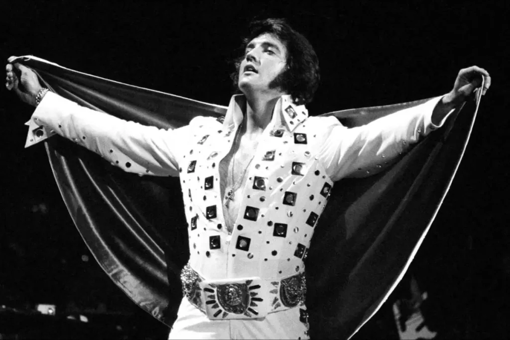 Black-and-white photo of Elves Presley in a studded white jumpsuit with arms outstretched during a dramatic stage moment.