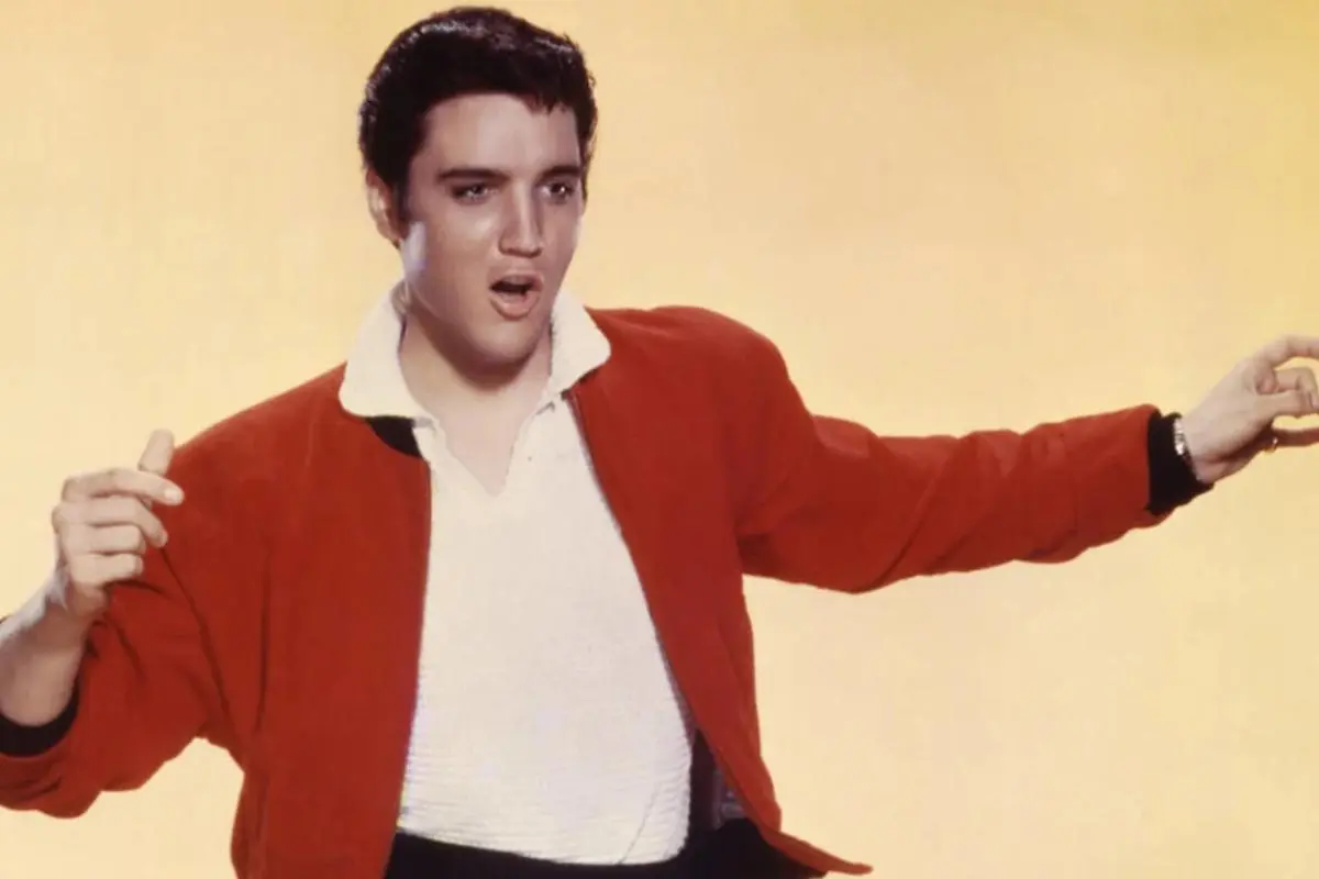 Elvis Presley  in a red jacket gestures mid-performance against a bright, minimal background.