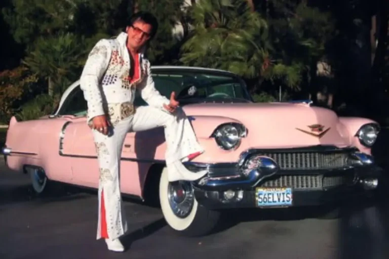 Elvis Presley dressed in a white, rhinestone-covered jumpsuit poses confidently beside a vintage pink Cadillac, resting one foot on the bumper and giving a playful hand gesture.