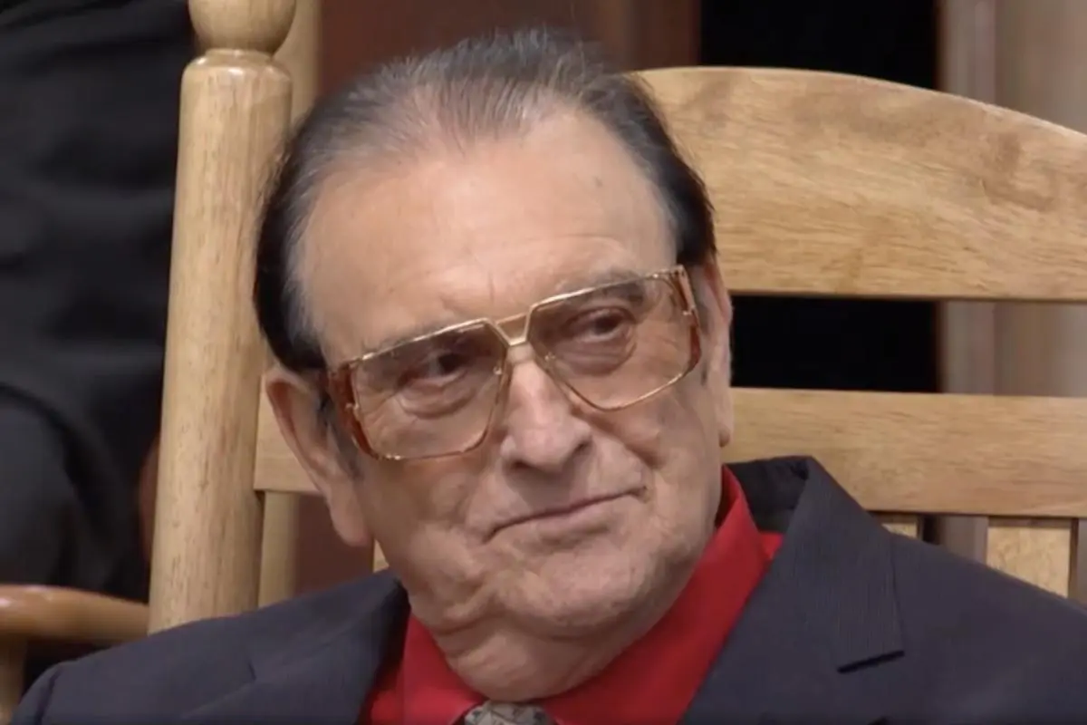 Ernest Tubb seated in a wooden chair, wearing glasses and a dark suit with a red shirt, looking thoughtfully off to the side.