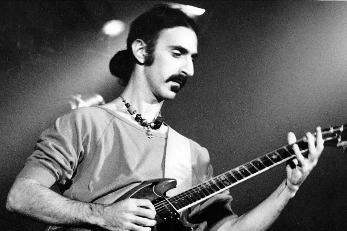 Frank Zappa long hair tied back, playing an electric guitar onstage under soft lighting.