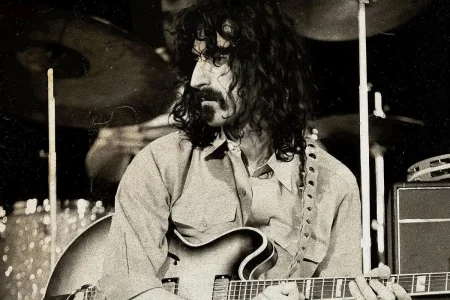 Frank Zappa with a mustache playing a semi-hollow electric guitar onstage, with drums visible in the background.