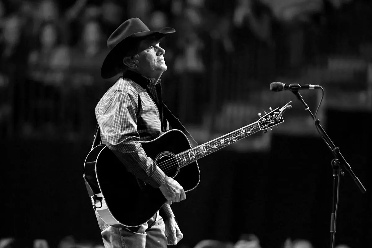 George Strait performing live, wearing a cowboy hat and holding an acoustic guitar on stage.