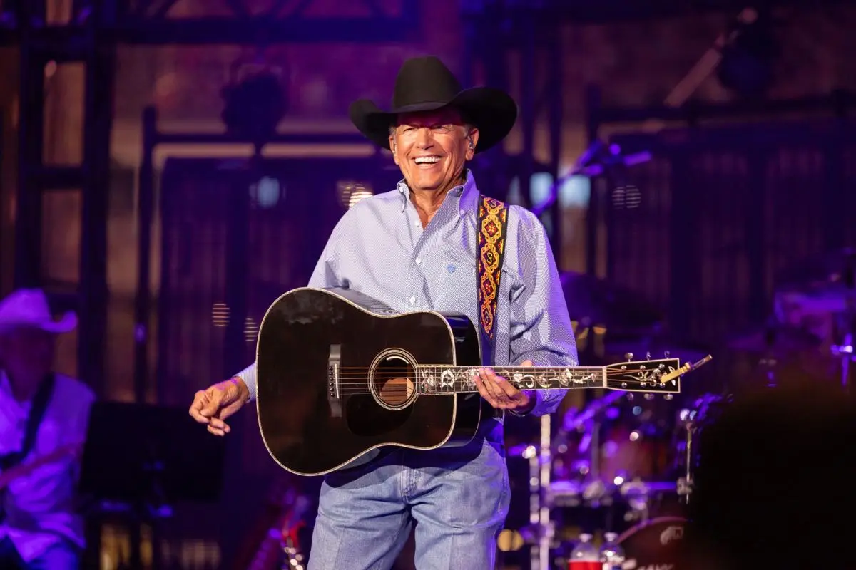George Strait smiling on stage, playing acoustic guitar under concert lights.
