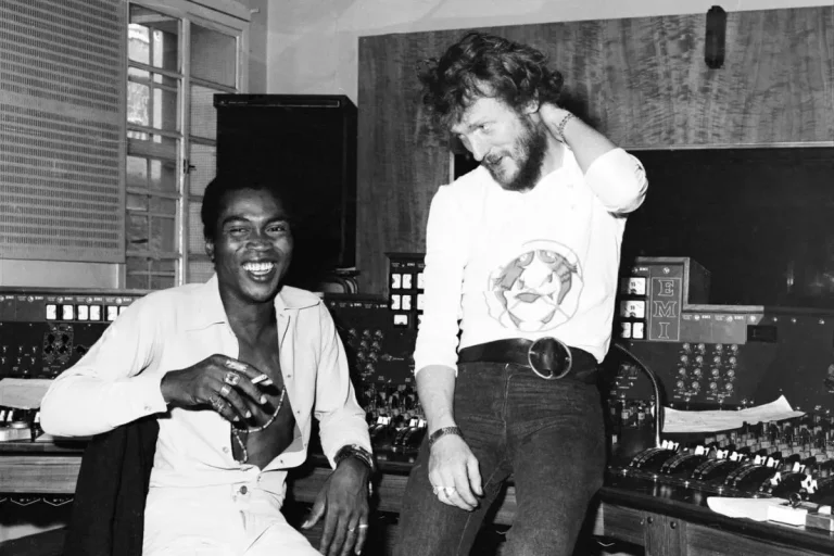 Ginger Baker and Fela Kuti sit together in a recording studio, smiling and relaxed, surrounded by audio equipment and mixing consoles.