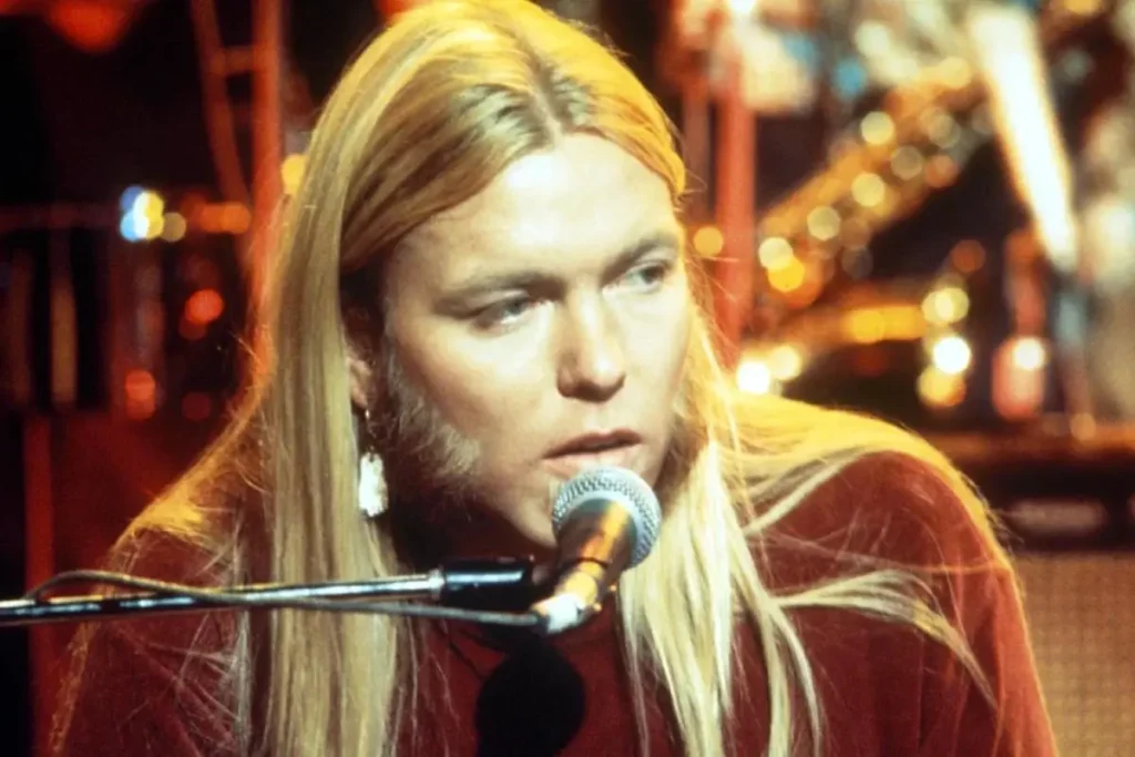 Gregg Allman, with long blond hair and sideburns, sings into a microphone while seated at a keyboard during a live performance.