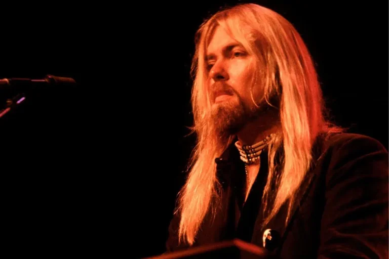 Gregg Allman performing on stage under warm red lighting, long blond hair and beard visible.
