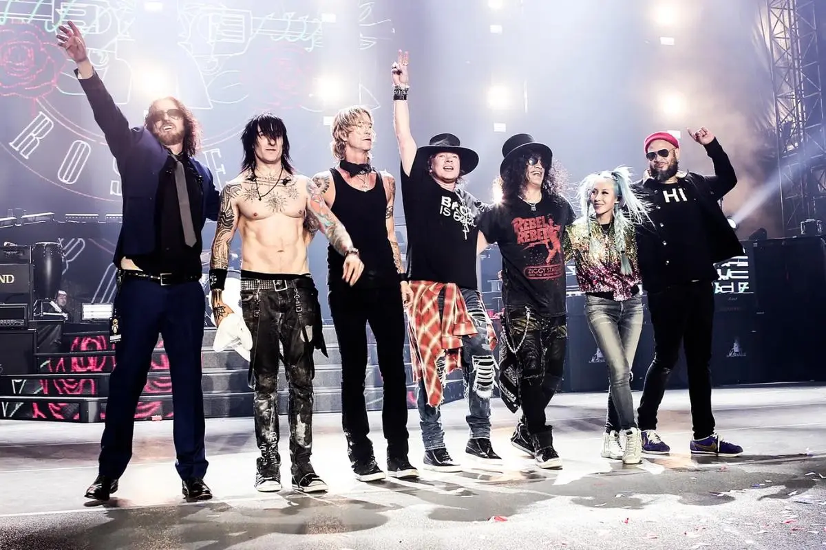 Guns N Roses Band standing together onstage after a concert, arms raised under bright stage lights.