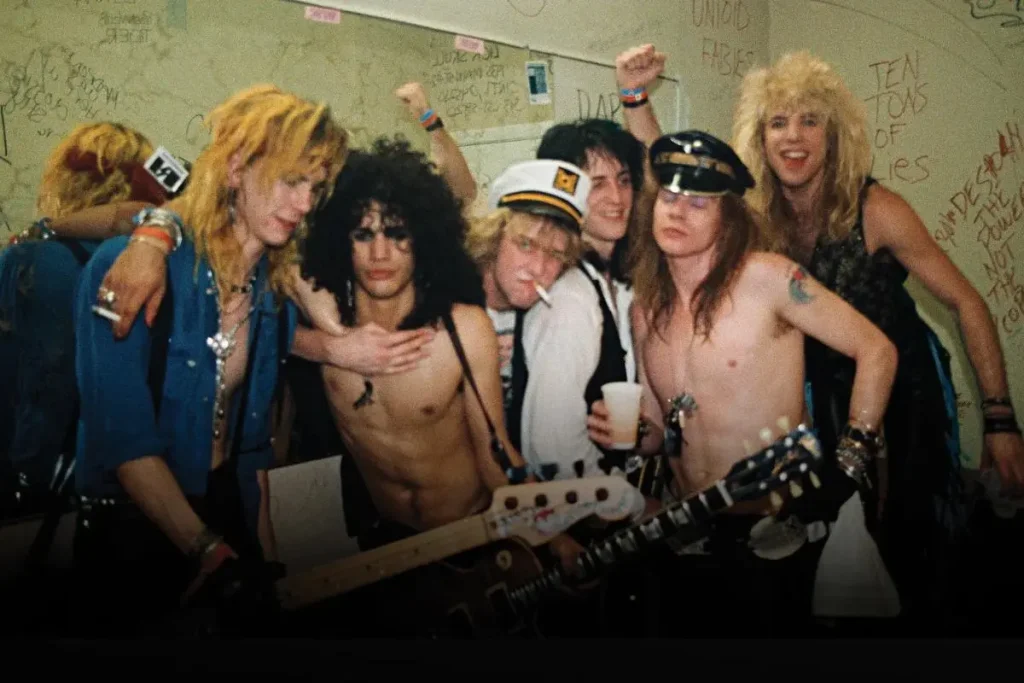Guns N Roses Band crowded together and smiling, wearing 1980s-style outfits and accessories.