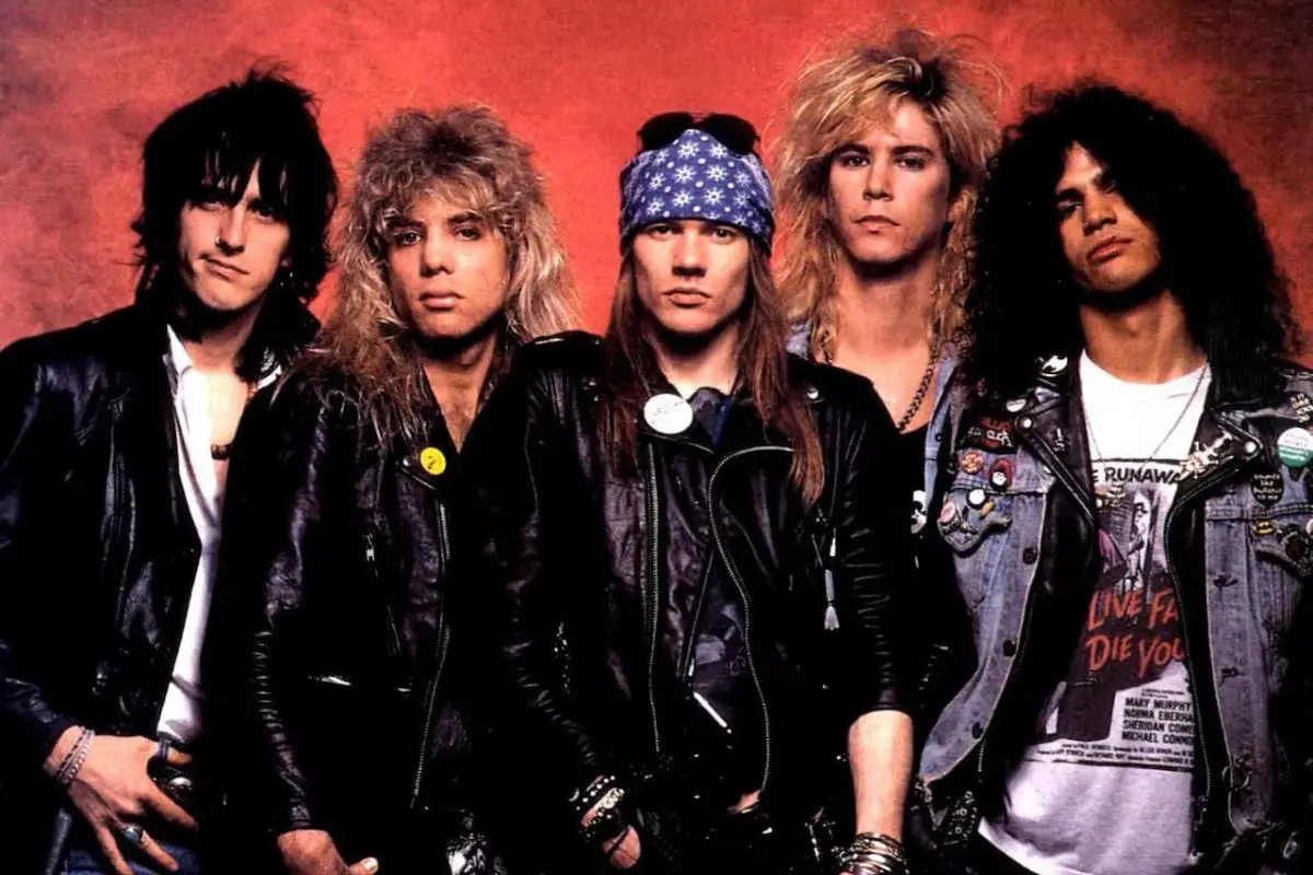 Studio portrait of Guns N Roses Band wearing leather jackets and bandanas, standing shoulder to shoulder against a red background.