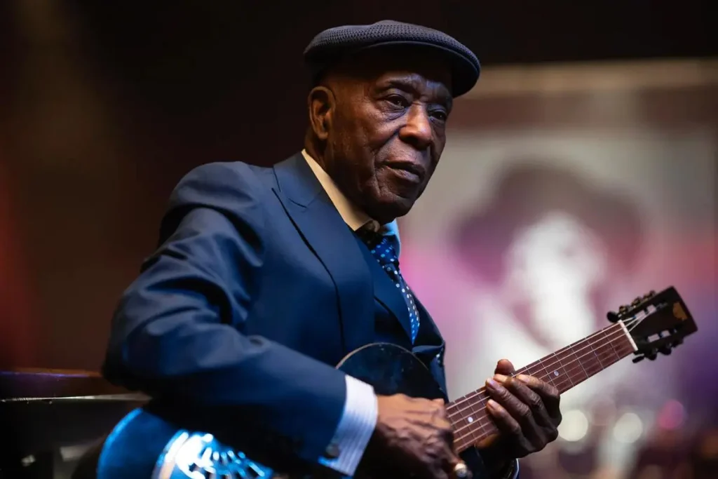 Buddy Guy's in a dark suit, dotted tie and flat cap, seated and cradling a dark archtop guitar.