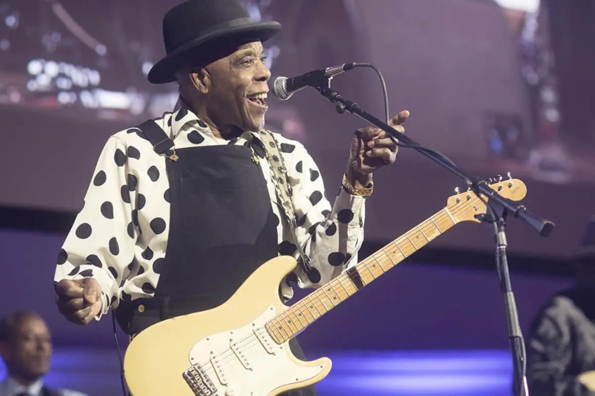 Buddy Guy's in a large-polka-dot shirt and black bib overalls, smiling and pointing while singing into a microphone.