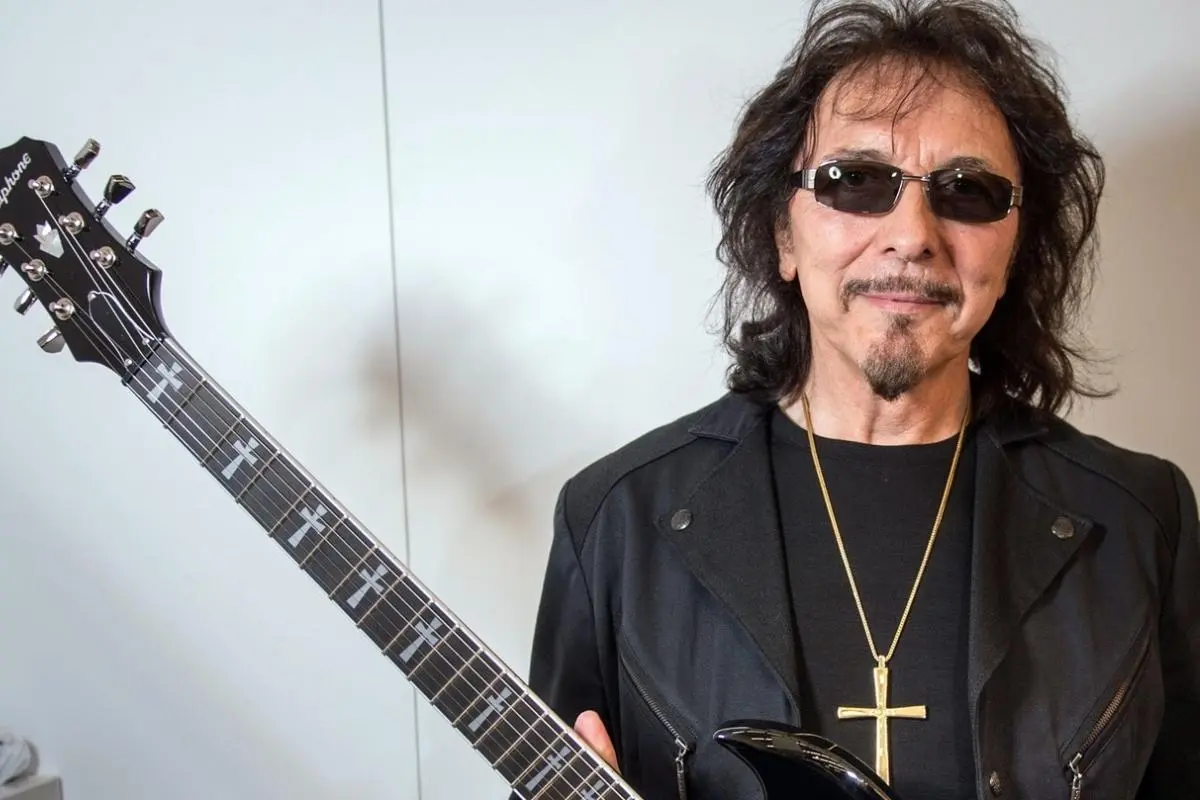 Tony Iommi posing indoors holding an electric guitar, wearing dark sunglasses, a black jacket, and a gold cross necklace, standing against a light-colored wall.