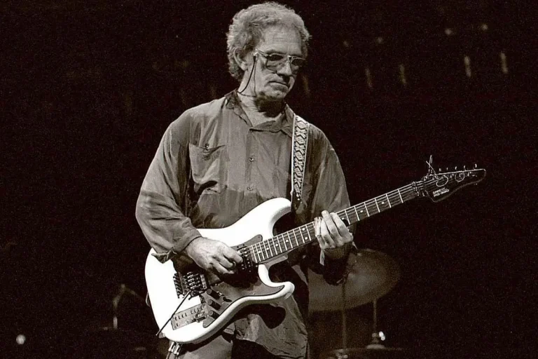 JJ Cale plays an electric guitar onstage, wearing glasses and a loose shirt, focused under dim concert lighting.