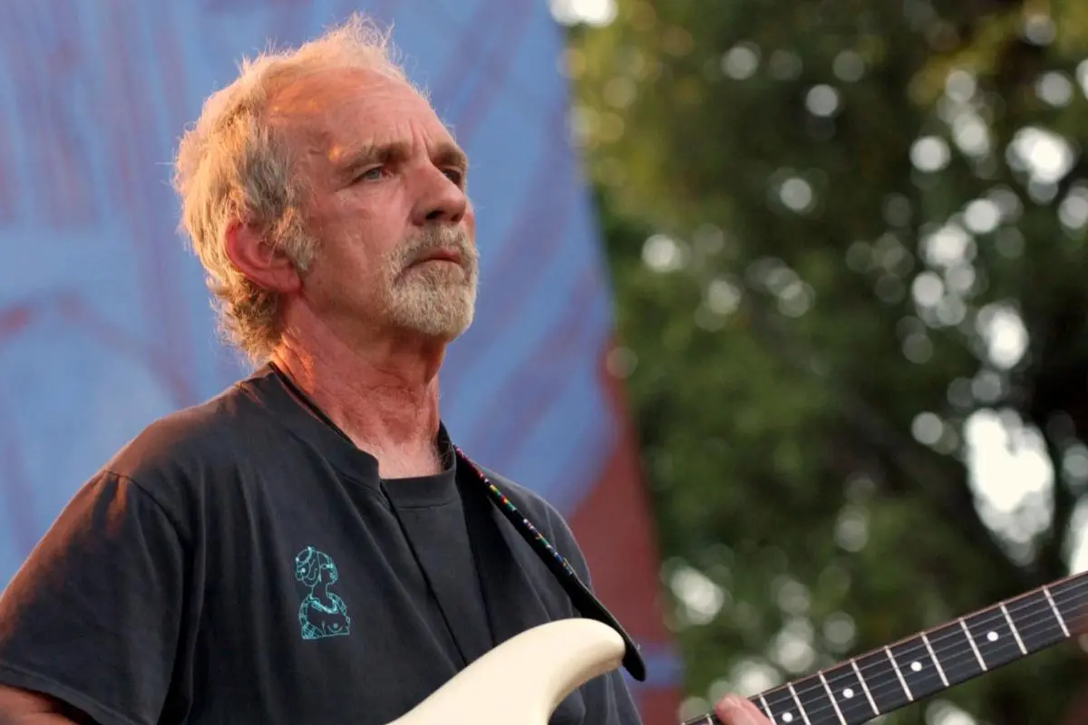 JJ Cale performs outdoors holding a white electric guitar, looking off into the distance with a calm, thoughtful expression.