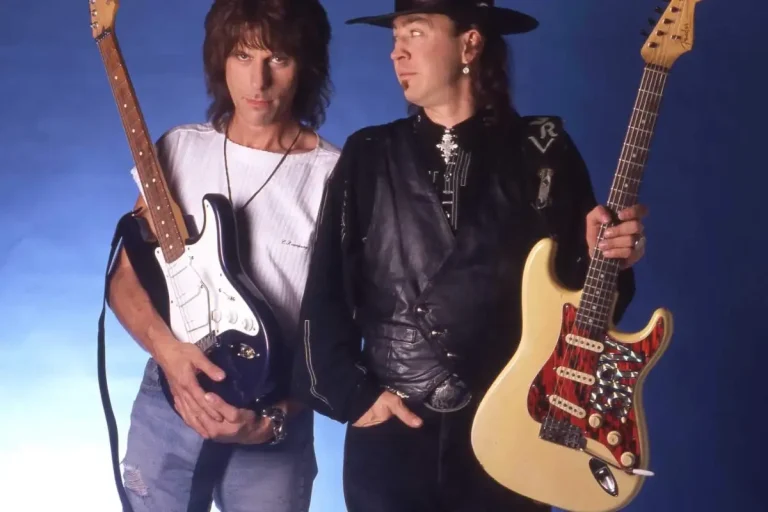 Jeff Beck and Stevie Ray Vaughan stand side by side holding their electric guitars, Jeff Beck looking directly at the camera while Stevie Ray glances toward him.