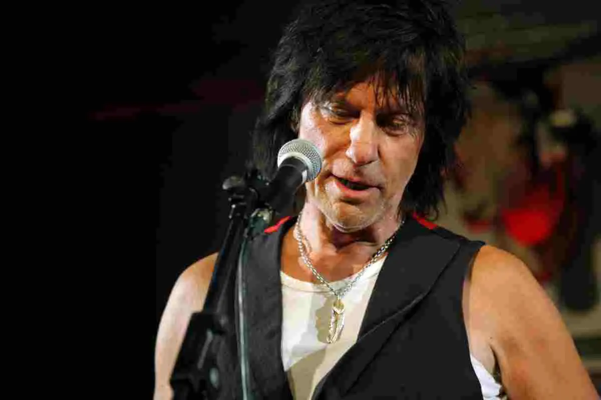 Jeff Beck performing on stage, singing into a microphone while wearing a black vest.