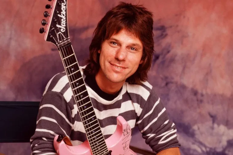 Jeff Beck smiling while holding a pink electric guitar in a studio portrait.