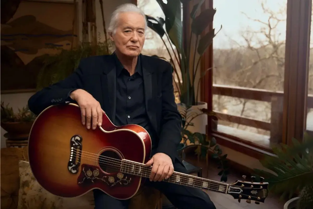Jimmy Pageseated indoors holding an acoustic guitar near a window.
