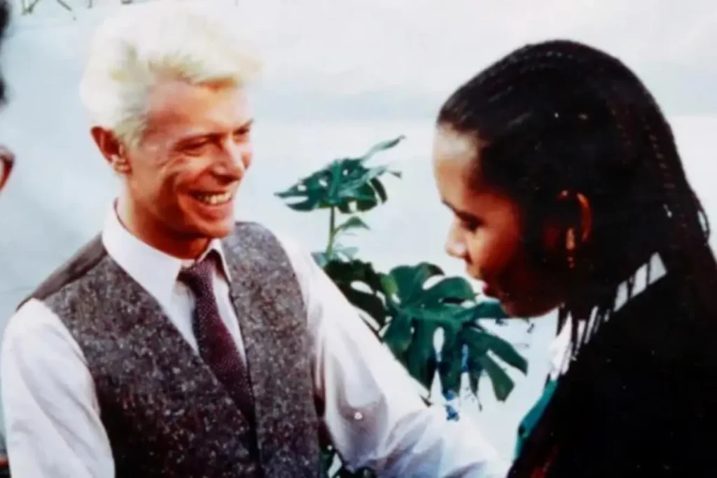 Joelene King and David Bowie share a warm, candid moment in conversation indoors.