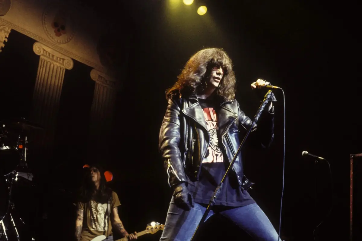 Joey Ramone performing on stage in a leather jacket and jeans, gripping a microphone stand and projecting raw energy and defiance during a live show.