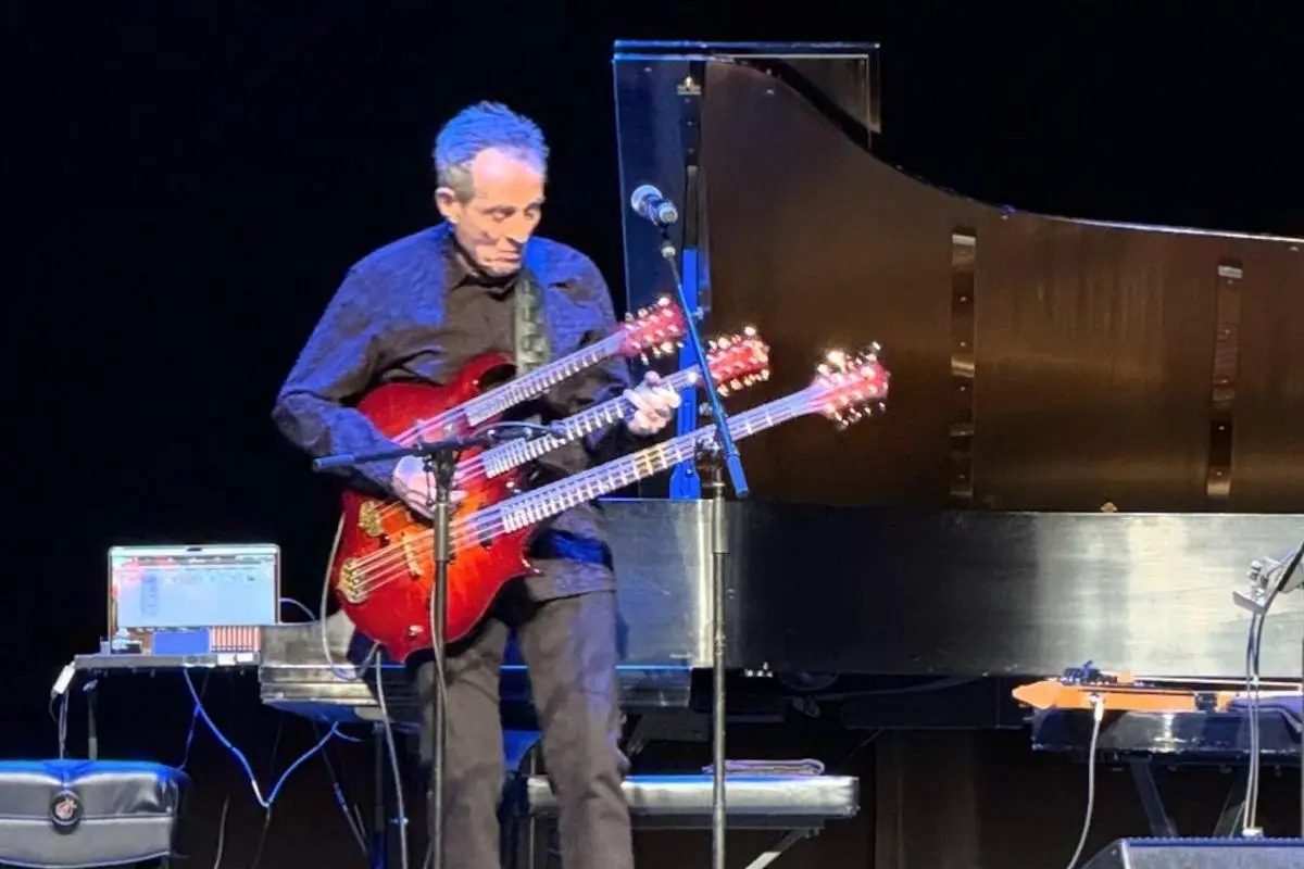John Paul Jones stands onstage playing a red double-neck guitar, with a piano visible behind him.