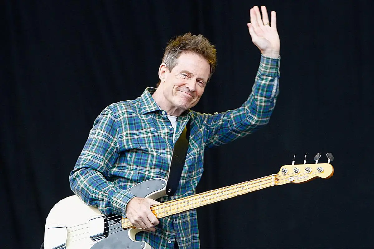 John Paul Jones waves to the audience while holding a white electric bass against a dark background.