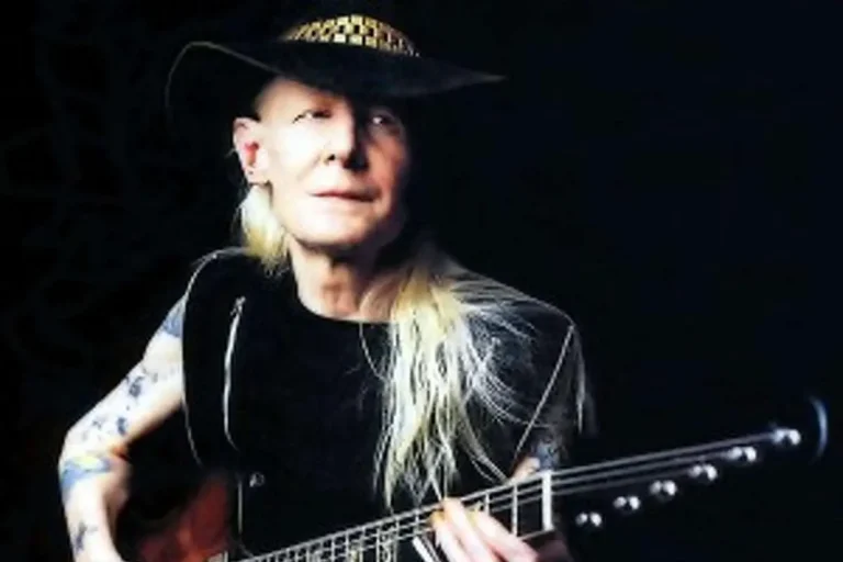 Johnny Winter’s Wild Climb From Pop Chaser To Slide Guitar Assassin Johnny Winter with long blond hair and tattoos plays guitar while wearing a wide-brimmed hat, illuminated against a dark background.