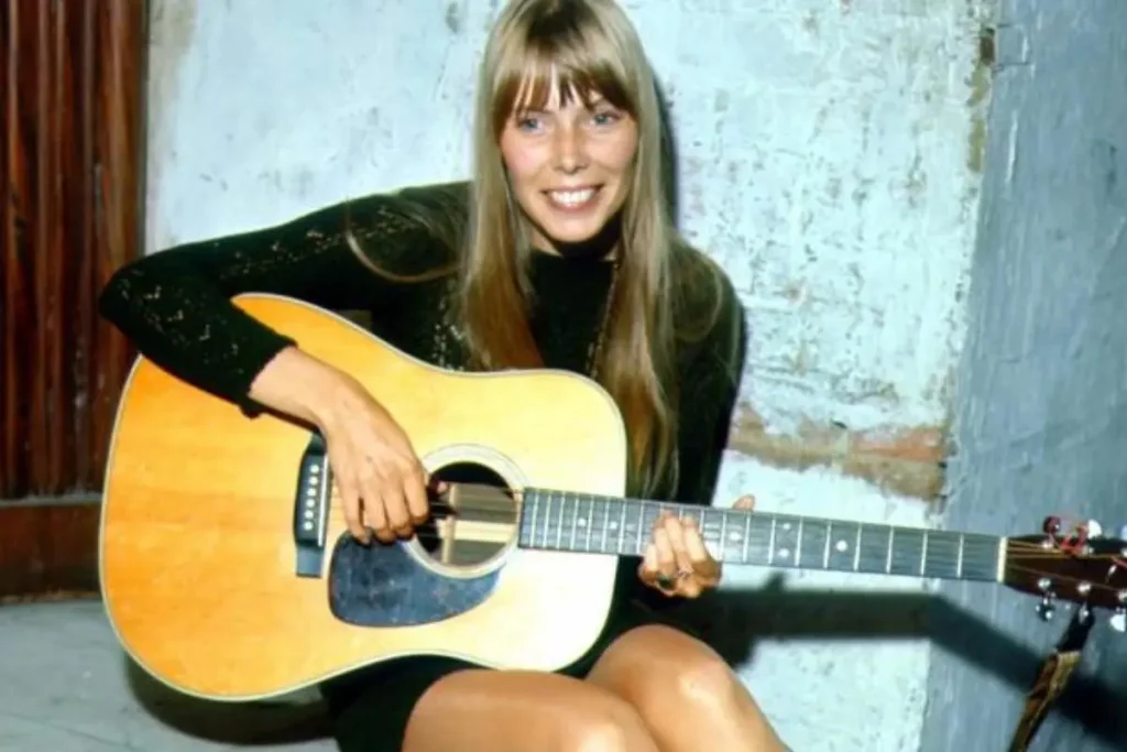 Joni Mitchell smiling while holding an acoustic guitar, seated indoors.