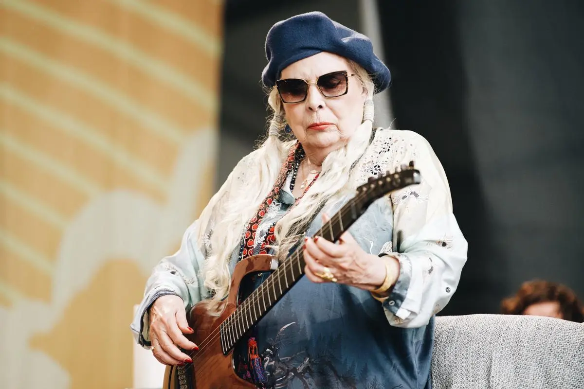 Joni Mitchell performing onstage, wearing a beret and sunglasses, playing guitar.