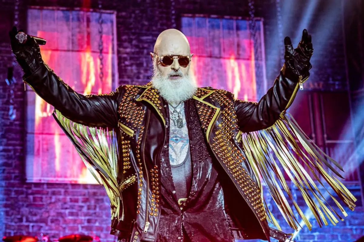 Judas Priest’s Rob Halford onstage wearing a studded leather jacket with fringes, arms raised under colorful stage lights.