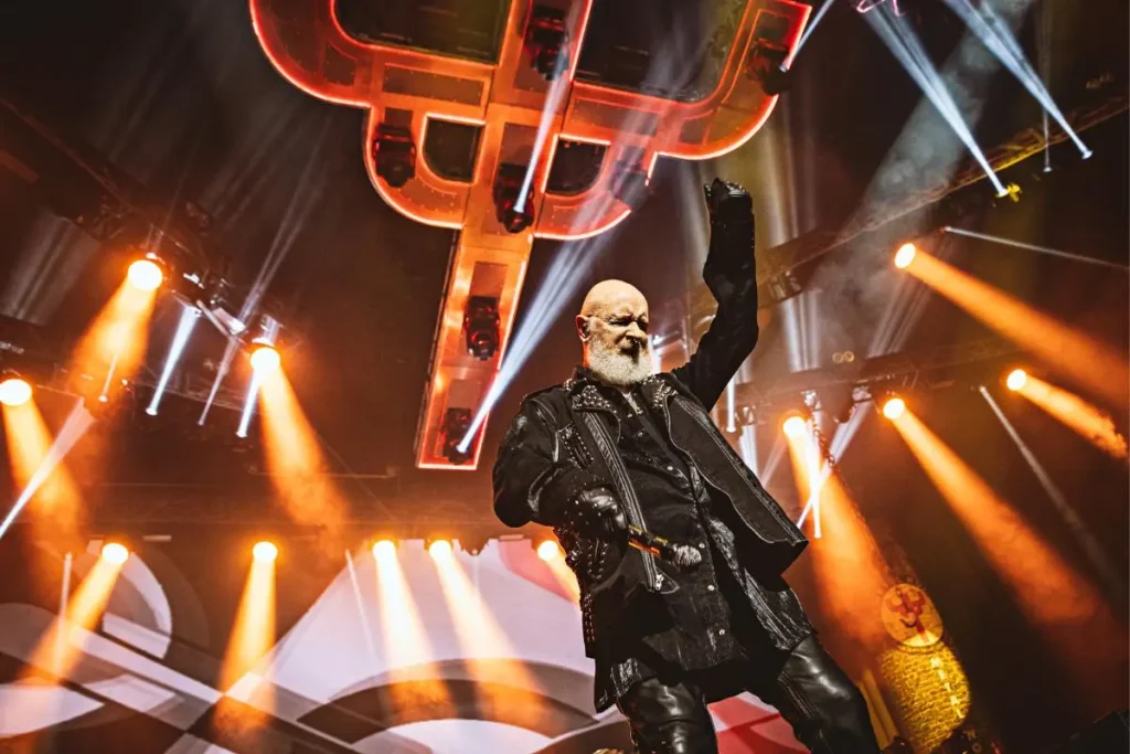 Rob Halford performing beneath a glowing Judas Priest cross, raising one arm amid dramatic concert lighting.