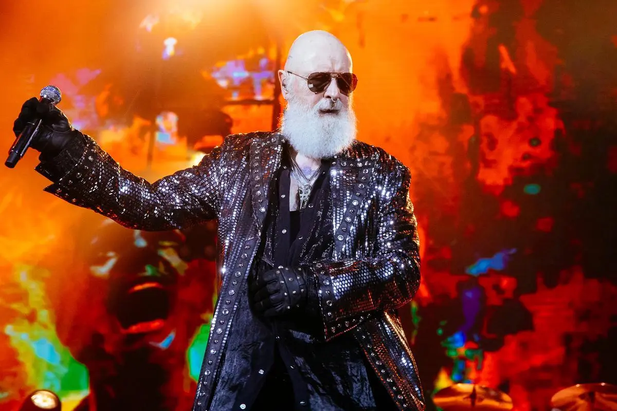 Rob Halford of Judas Priest performing live, holding a microphone against a fiery orange stage backdrop.