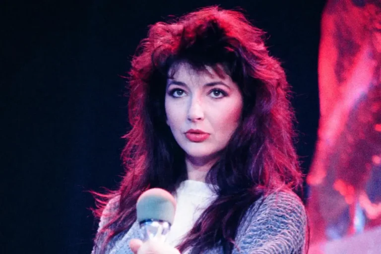Kate Bush holding a microphone onstage under pink and purple lighting.