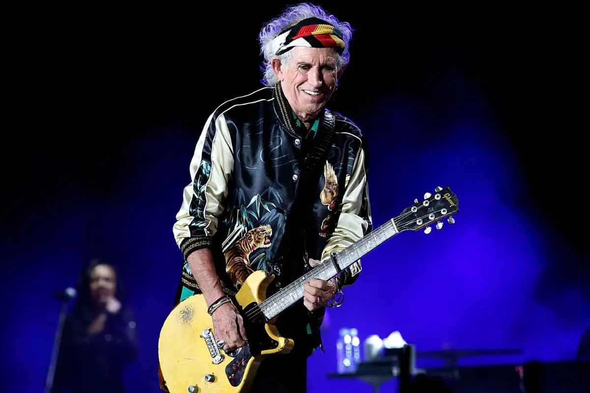 Keith Richards smiling while playing an electric guitar under blue stage lights.
