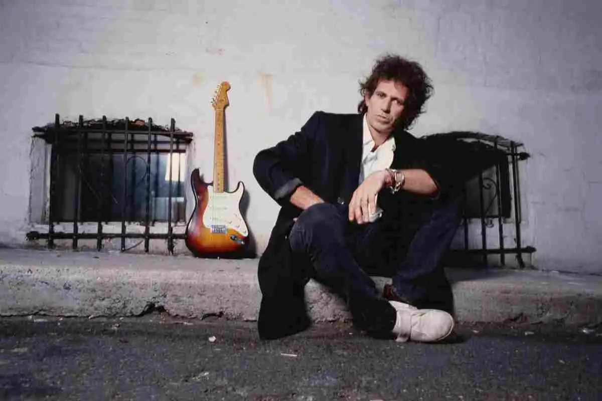 Keith Richards sitting on steps beside an electric guitar, wearing a dark jacket and jeans.