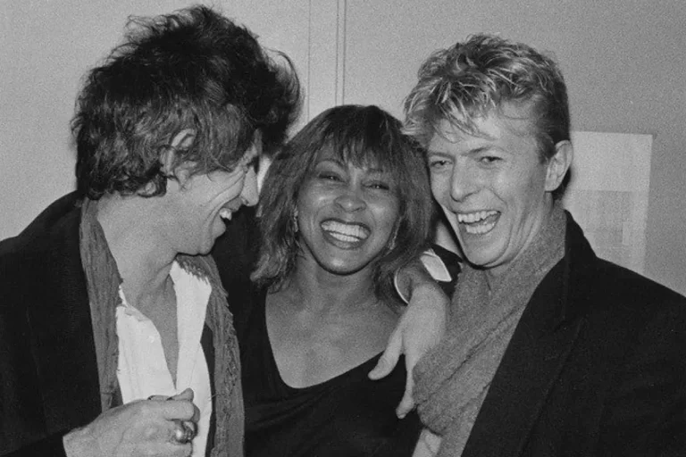 Keith Richards, Tina Turner and David Bowie share a joyful backstage moment, laughing with their arms around one another.