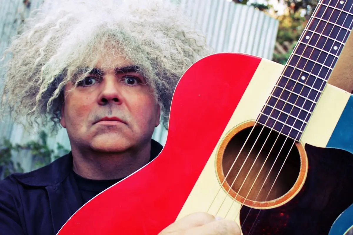 Portrait of King Buzzo with wild, curly gray hair holding a brightly colored acoustic guitar, conveying individuality and unconventional artistic style.