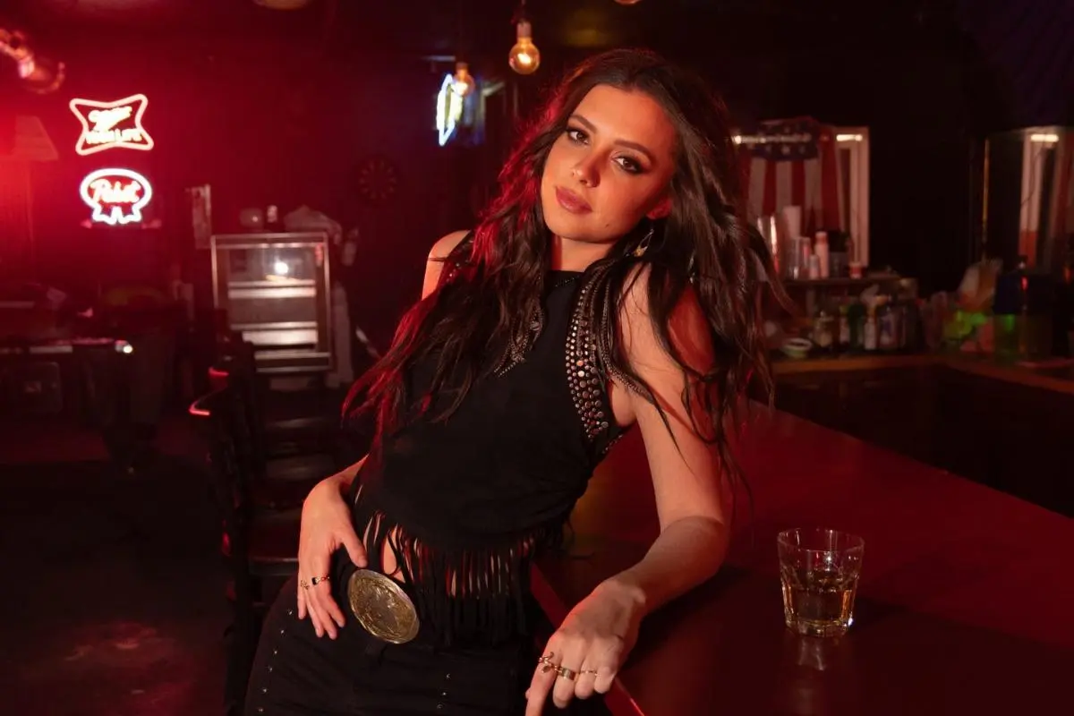 Portrait of Lanie Gardner leaning against a bar under warm red lighting, wearing a black outfit with a statement belt.