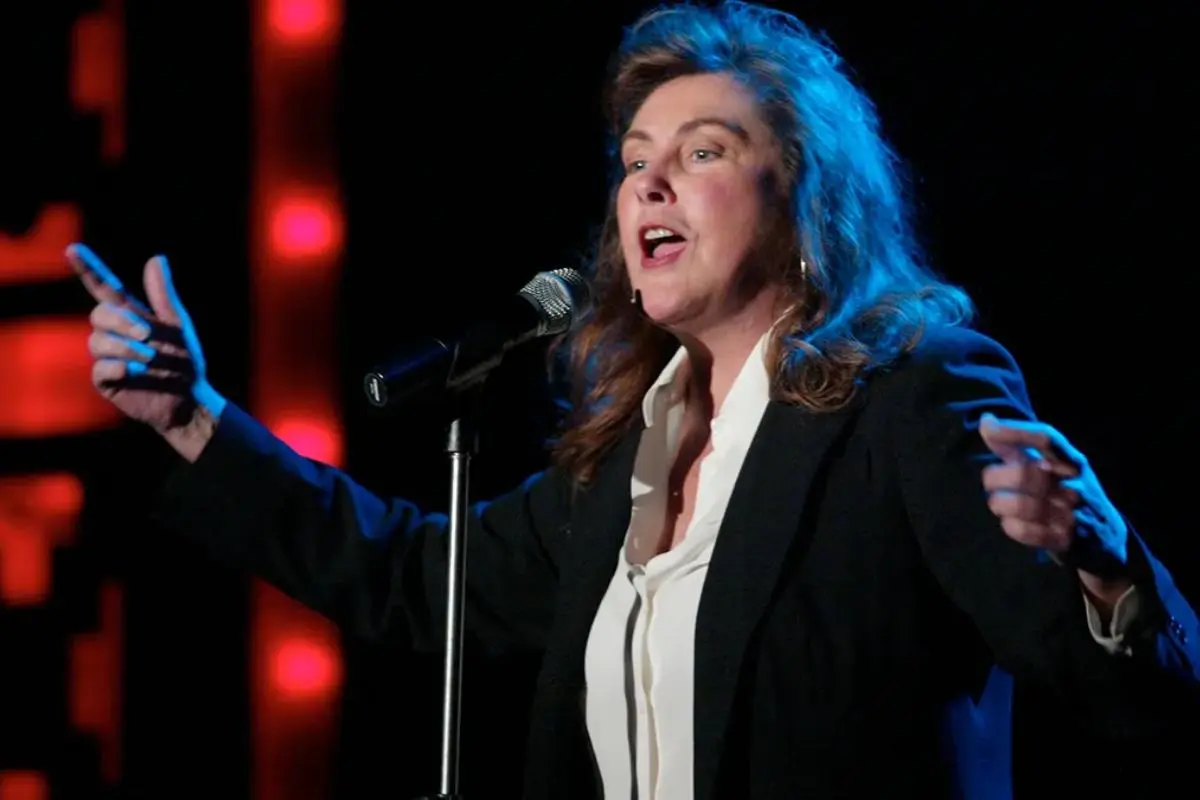 Laura Branigan performing on stage, singing into a microphone with arms extended, wearing a black blazer and white blouse under dramatic concert lighting.
