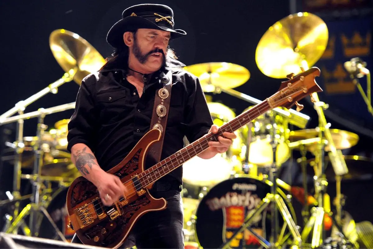Lemmy Kilmister wearing a black hat plays an electric bass onstage, with bright stage lights and a drum kit behind him.