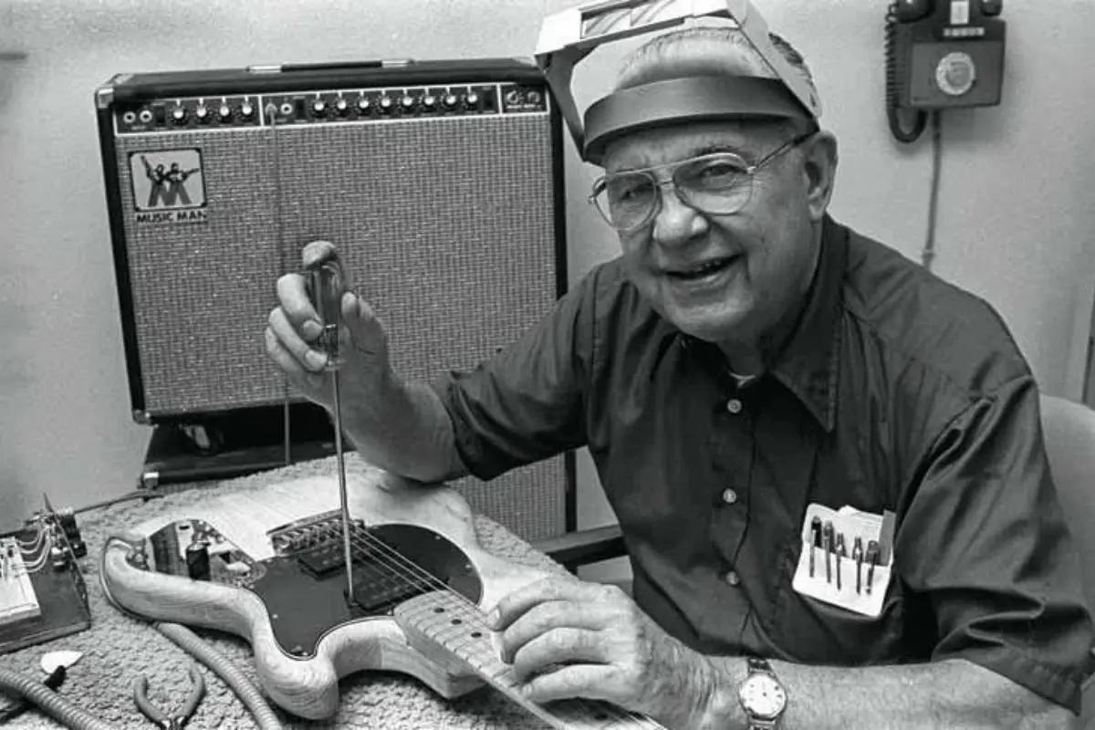 Leo Fender smiling beside a Fender amplifier, holding a guitar tool.