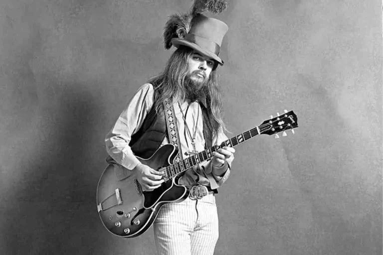 Leon Russell posing in a black-and-white studio photo while playing an electric guitar, wearing a tall top hat with a feather, long hair, a beard, and vintage-style clothing.