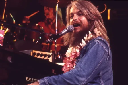 Leon Russell wearing a flower lei sings into a microphone while playing piano onstage.