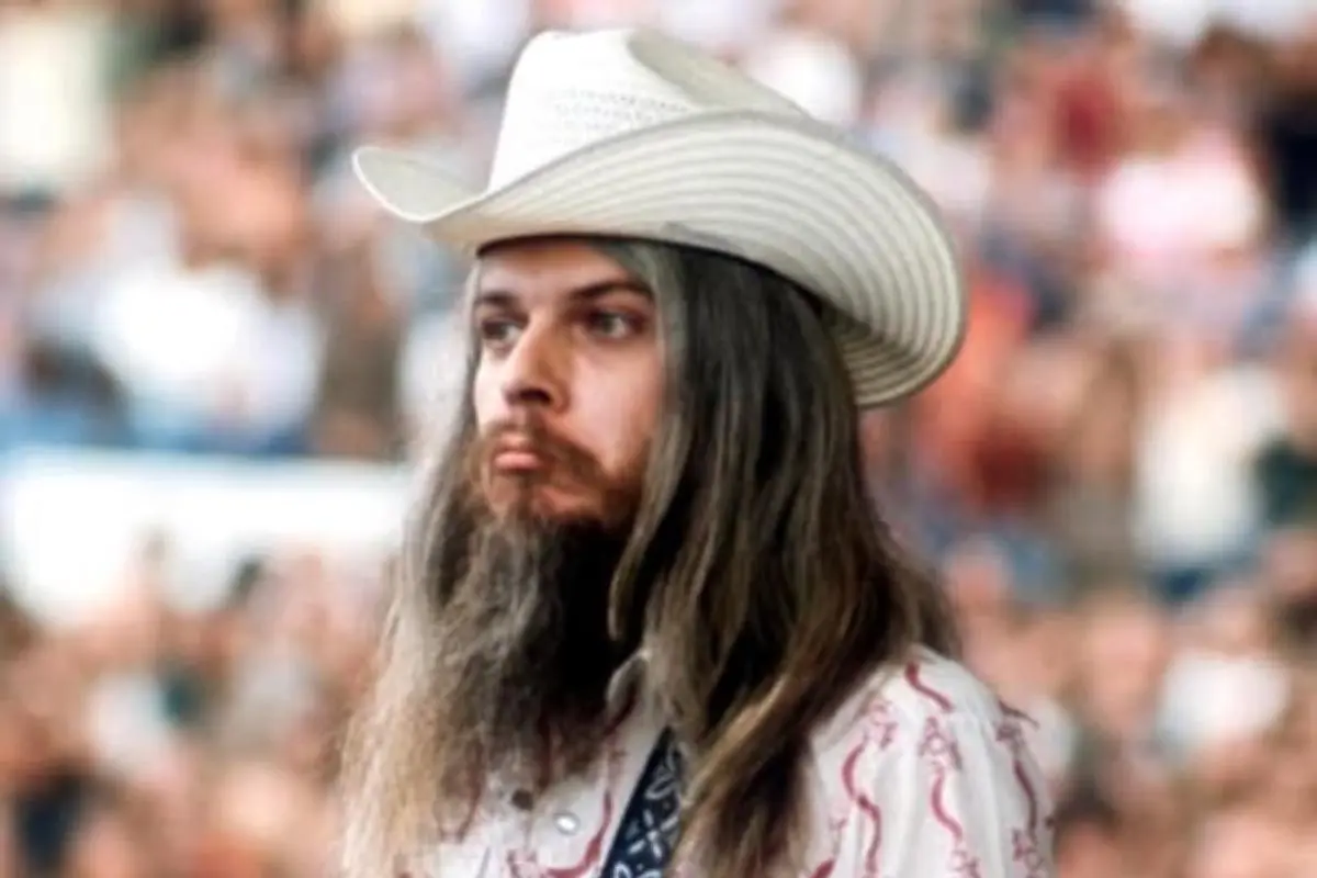 Leon Russell wearing a white cowboy hat and long hair, standing before a blurred crowd at an outdoor event.