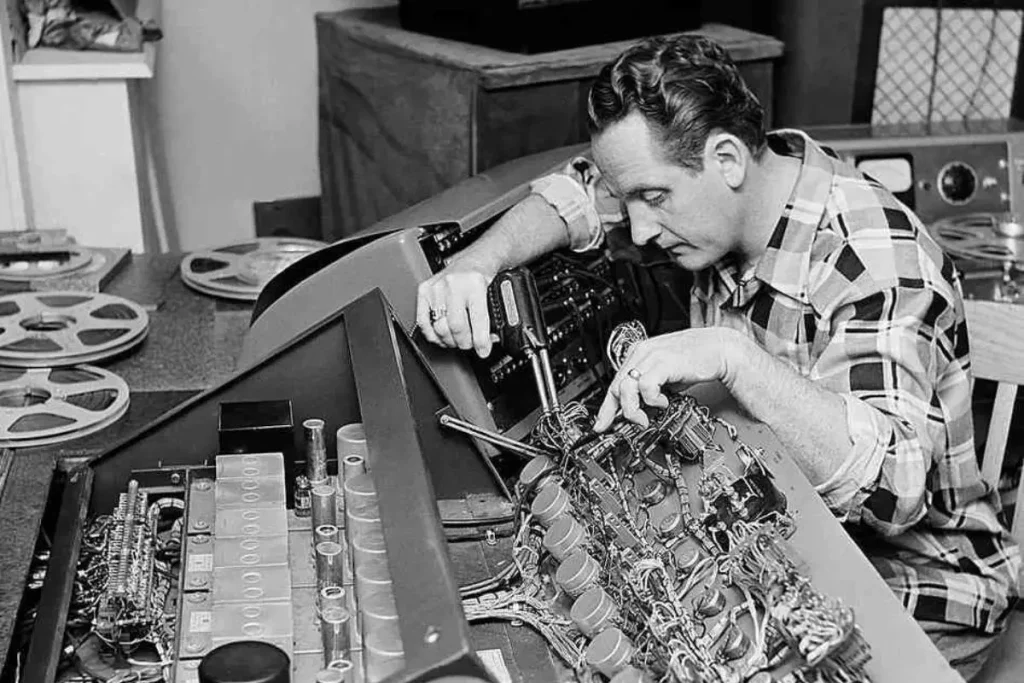 Les Paul, wearing a plaid shirt, leans over an open reel-to-reel recording machine, using tools to adjust the intricate wiring and components inside.