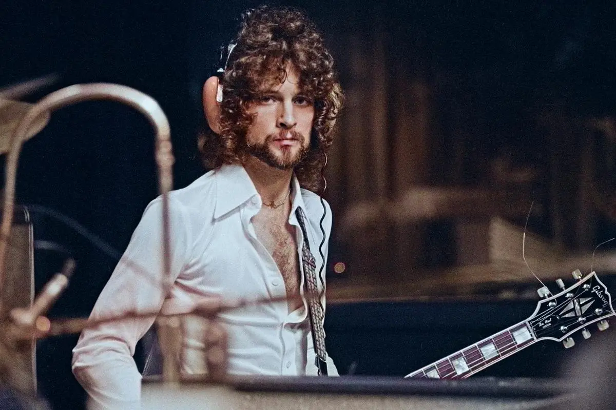 Lindsey Buckingham performing on stage, wearing a white open-collar shirt and playing electric guitar, with curly hair and a focused expression under warm stage lighting.