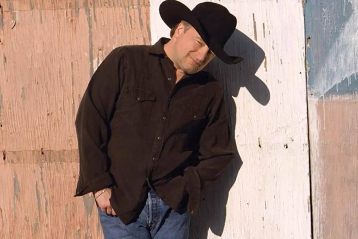 Mark Chesnutt leaning against a weathered wall, wearing a black cowboy hat, dark shirt, and jeans, posed casually with one hand in his pocket.